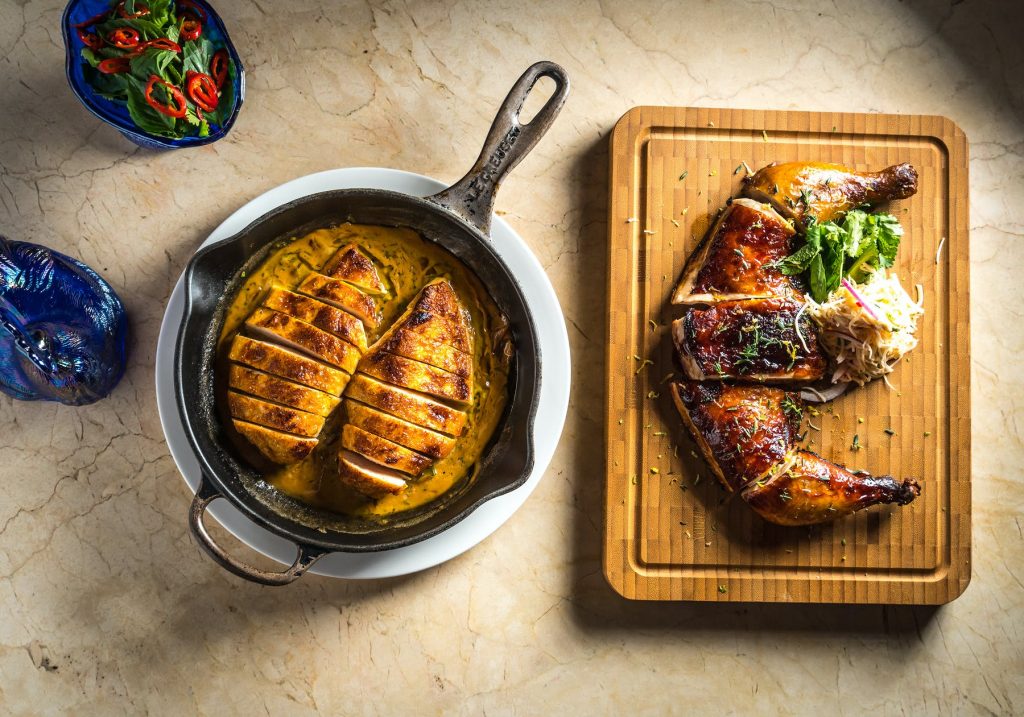 looking down onto two meals, one in a silver dish and one on wooden bread board with two blue side dishes 